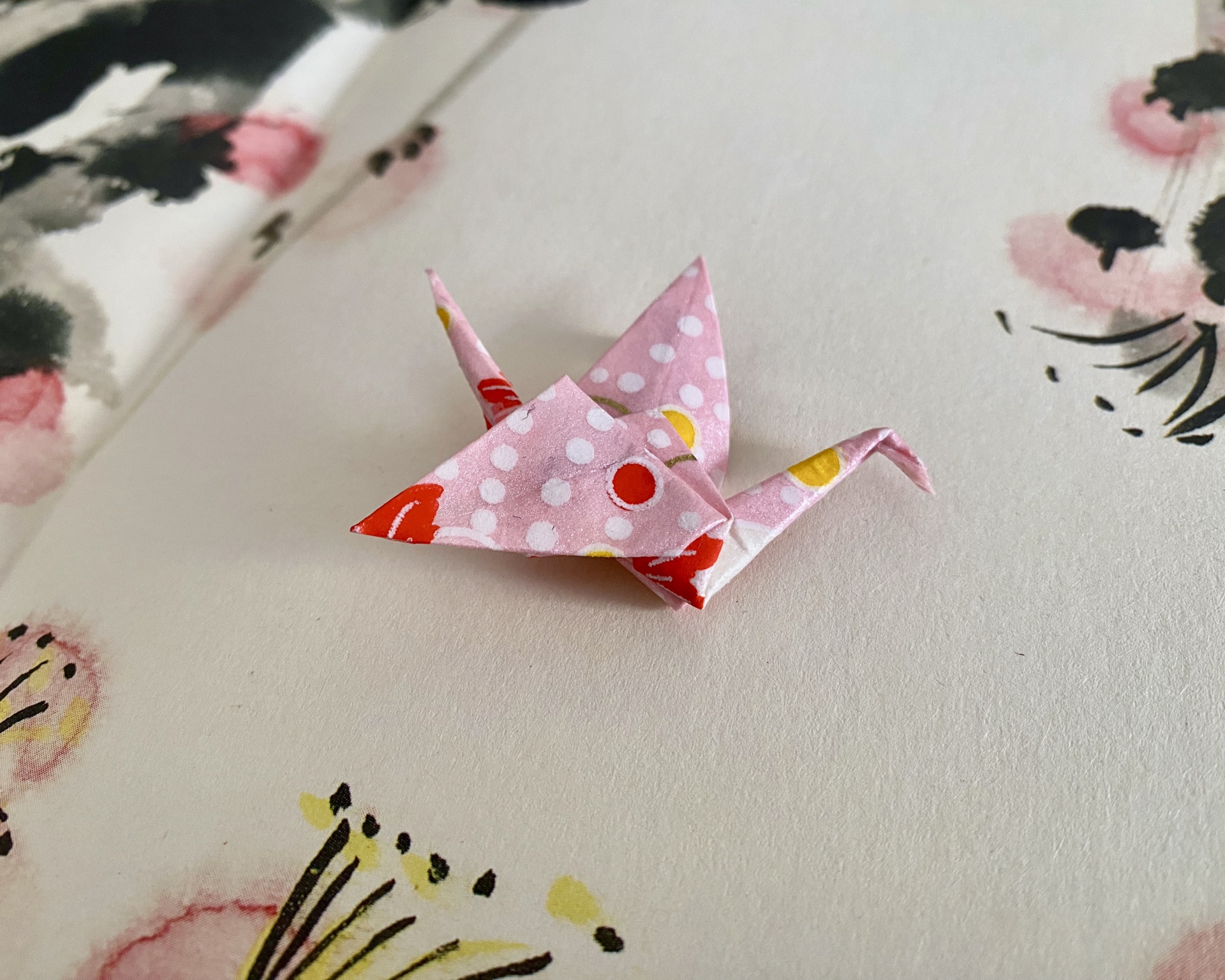 Pregadeira Tsuru Pequeno origami rosa com bolinhas e círculos, sobre papel branco com pintas florais