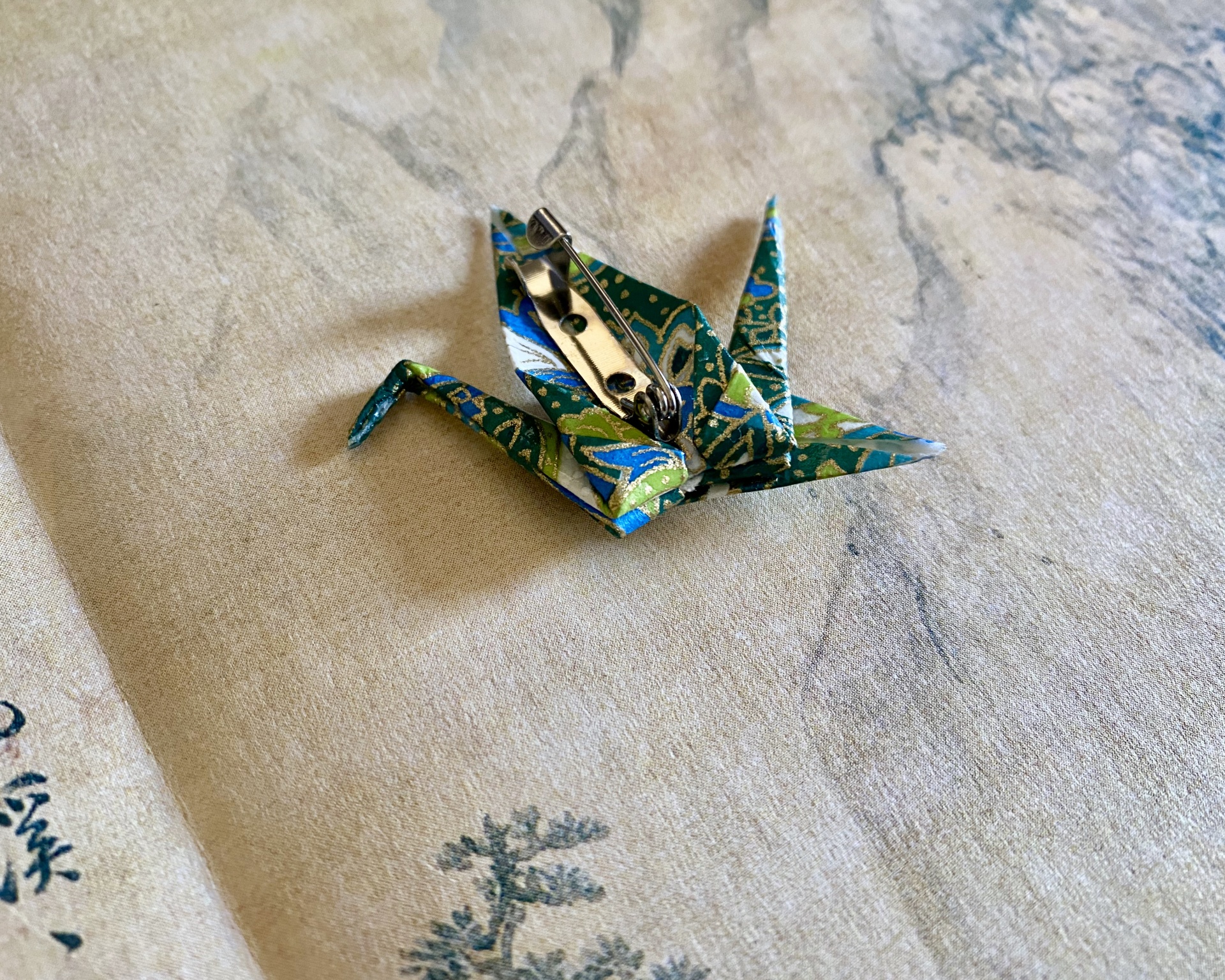 Pregadeira Tsuru Broche em papel origami colorido com base metálica numa superfície texturada.