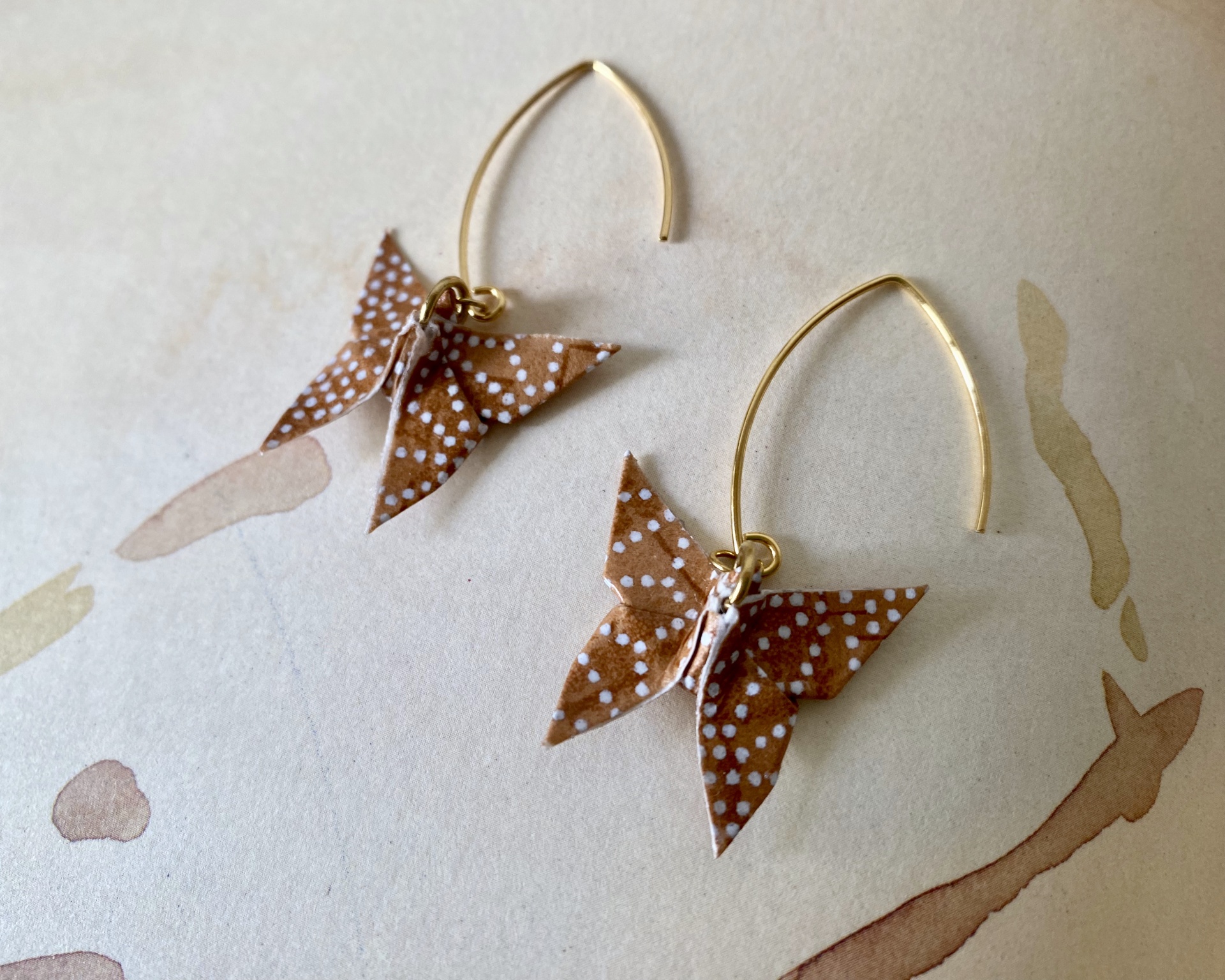 Brincos Borboleta Brincos de borboletas em papel castanho com pontos brancos e argolas douradas sobre fundo claro.