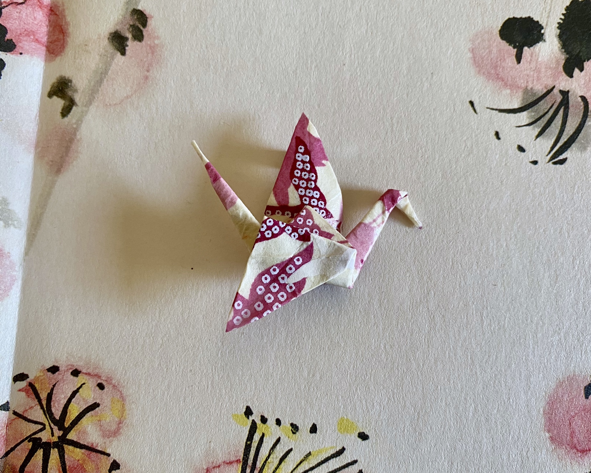 Pregadeira Tsuru Origami de pássaro em papel rosa e creme com padrão circular sobre fundo com padrão floral.