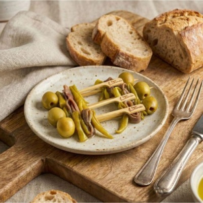 Prato com petiscos de azeitonas, pimentos verdes e anchovas, pão e talheres sobre mesa de madeira