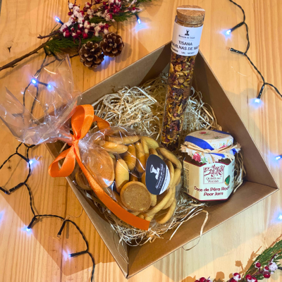 Conjunto de presente com biscoitos, tisana e compota numa caixa de cartão decorada com luzes, pinhas e flores secas.