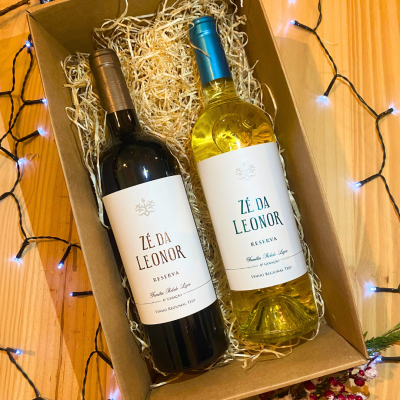 Duas garrafas de vinho ZÉ DA LEONOR RESERVA numa caixa de madeira com palha, sobre mesa de madeira com luzes decorativas brancas.