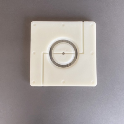 Peça quadrada branca com anel metálico circular no centro sobre fundo cinza