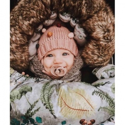 Bebé sorridente com gorro rosa e chupeta num casaco ou manta com padrão de folhas verdes e castanhas.