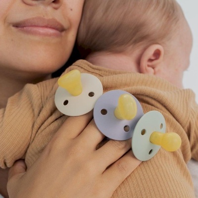 Três chuchas coloridas em tons pastel sobre a mão de mulher que segura um bebé vestido de malha camel