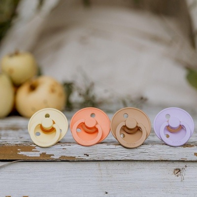 Quatro chupetas de silicone coloridas sobre madeira envelhecida com maçãs amarelas ao fundo