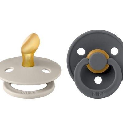 Duas chupetas BIBS, uma bege com bico dourado e outra cinzenta com detalhes dourados