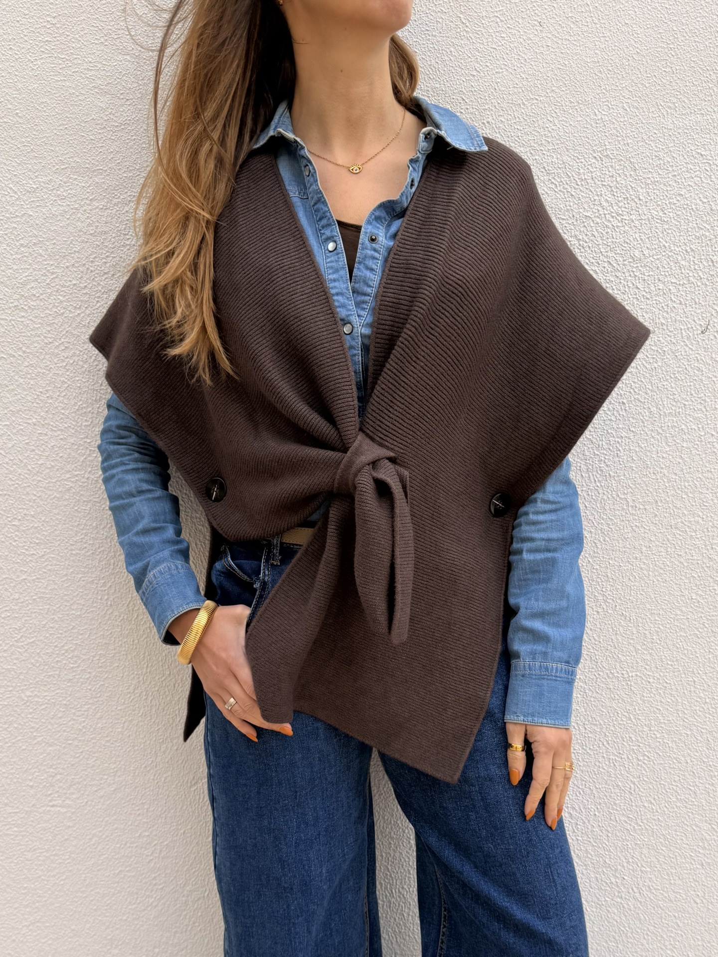 Mulher com colete de malha castanho, camisa de ganga azul e jeans azuis contra parede branca