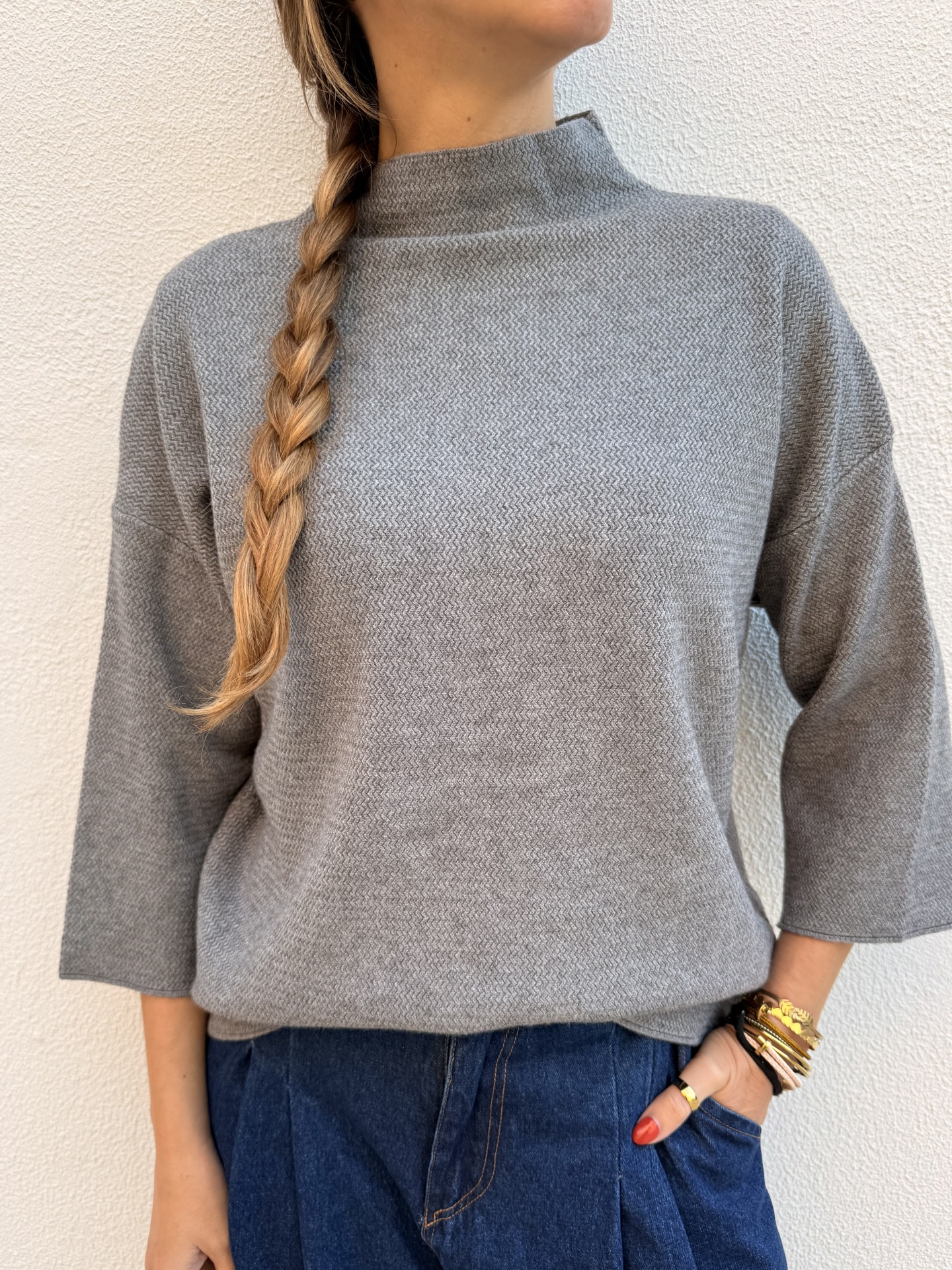 Sweater cinzento com gola subida e mangas 3/4, calças jeans azuis e pulseiras douradas, fundo branco.