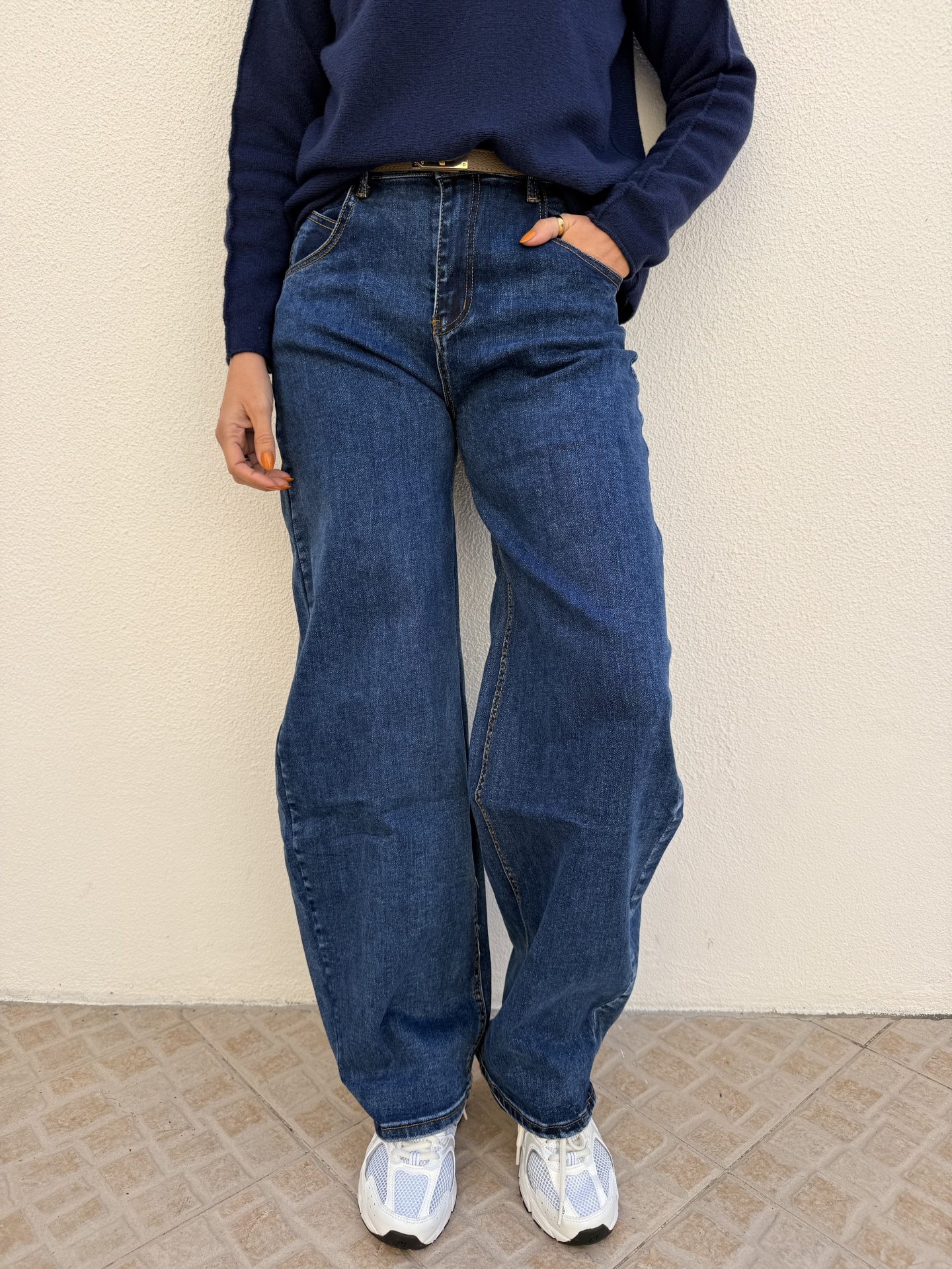 Pessoa vestindo calças de ganga largas azuis, camisola azul escura e ténis brancos em fundo de parede clara e chão de azulejo.