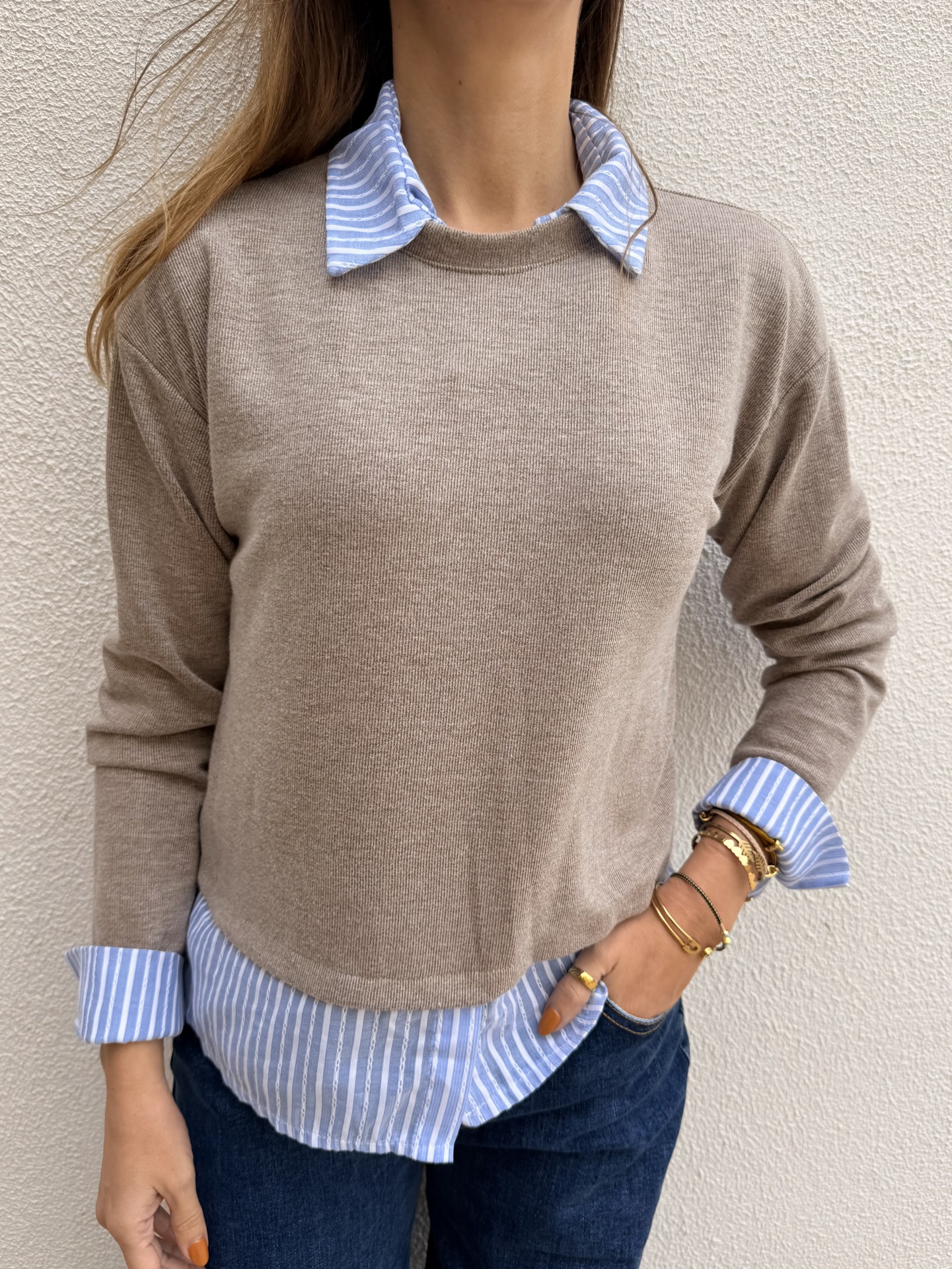 Pessoa com sweater castanho bege sobre camisa azul claro às riscas e calças de ganga azul escuro