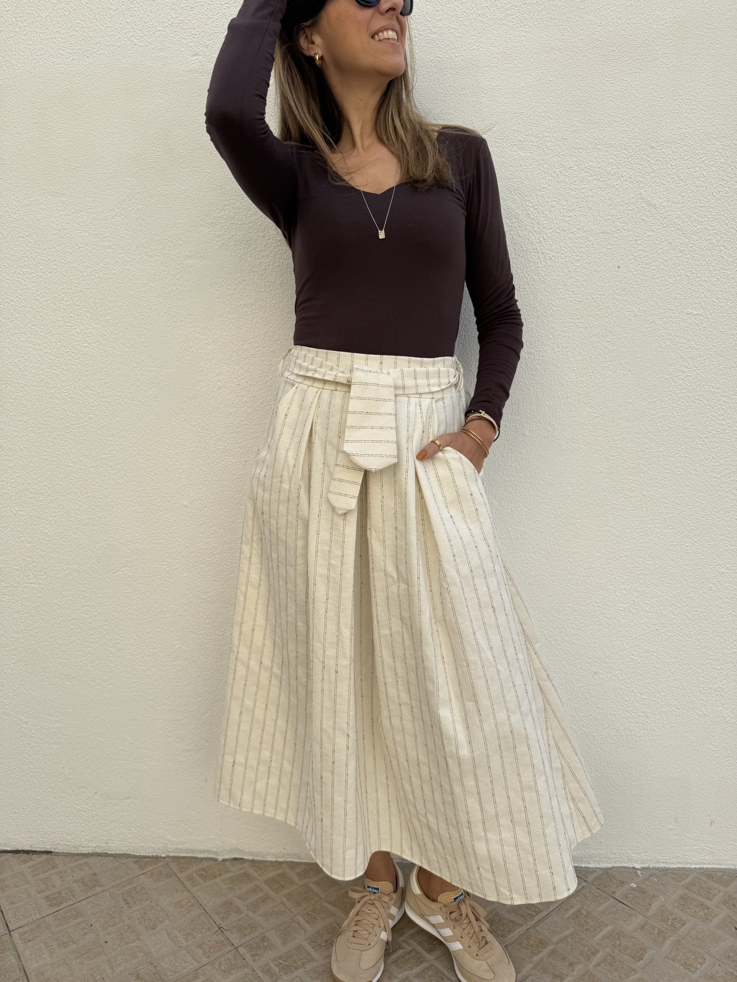 Mulher com blusa castanha, saia creme riscada e ténis beges junto a parede creme.