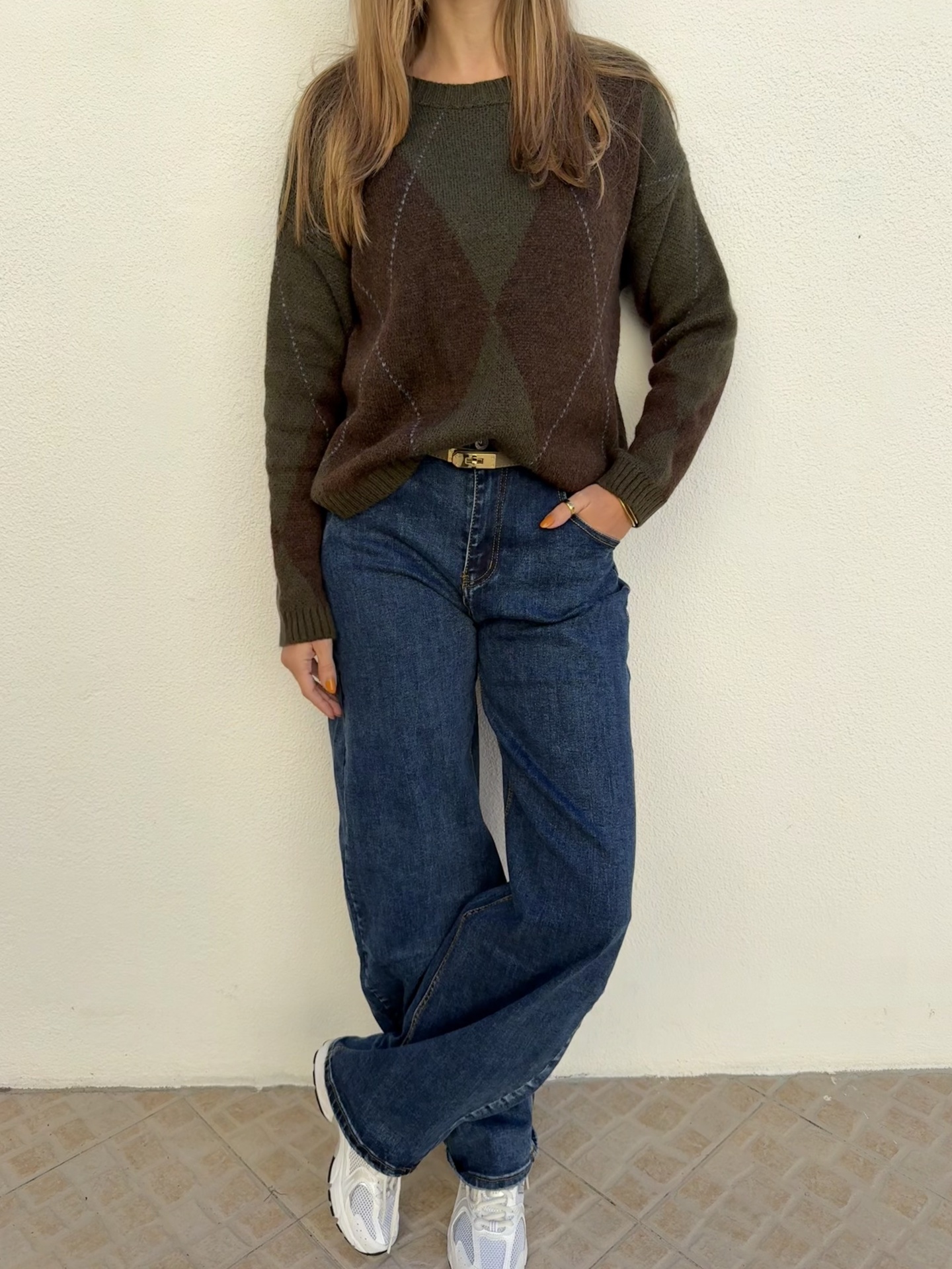 Pessoa a usar camisola de malha castanha de losangos, calças de ganga azul escuras largas e ténis brancos.