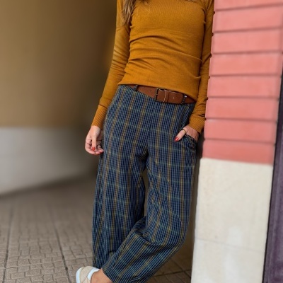 Pessoa vestindo camisola castanha, calças às riscas largas e ténis bege, encostada a parede de tijolos vermelhos.