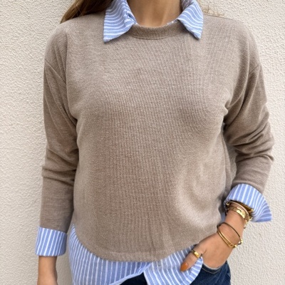 Pessoa com camisola bege sobre camisa azul riscada junto a parede clara