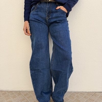 Pessoa vestindo calças de ganga largas azuis, camisola azul escura e ténis brancos em fundo de parede clara e chão de azulejo.