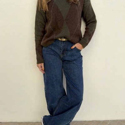 Pessoa vestindo camisola castanha e verde com padrão losango, calças de ganga azuis largas e ténis brancos, contra parede branca.