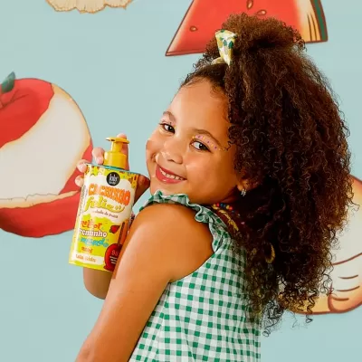 Criança sorridente com vestido xadrez verde e branca segurando frasco de cuidado capilar infantil