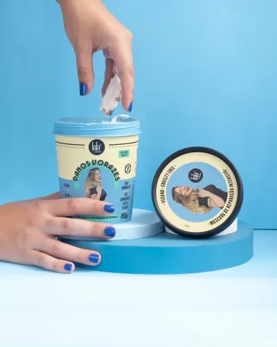 Dois frascos de máscara capilar Lola Cosmetics em fundo azul, um aberto com produto sendo retirado da embalagem.