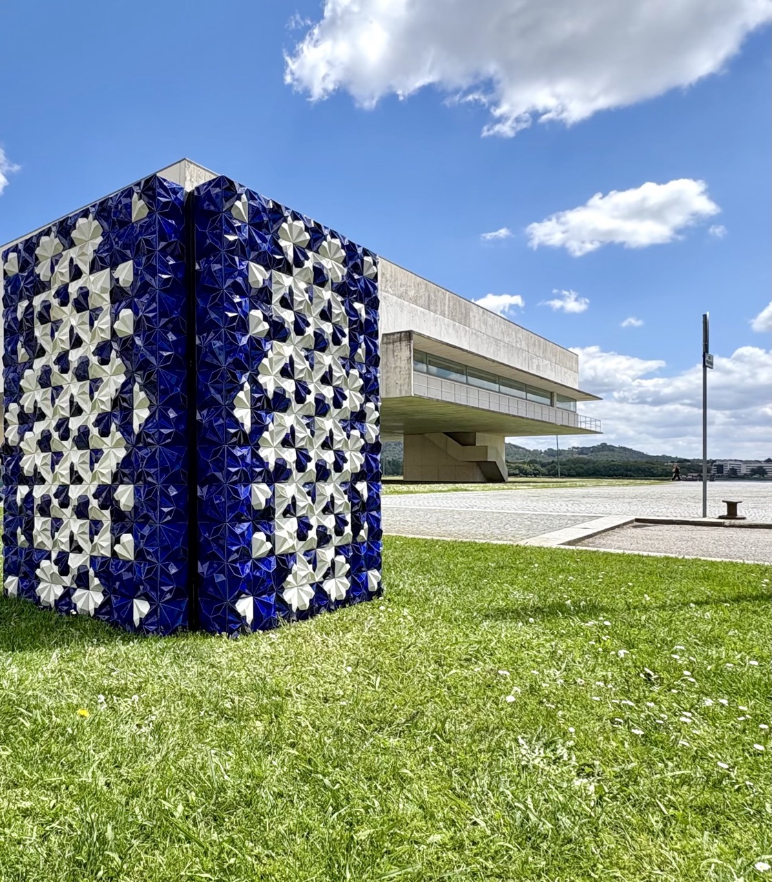 Tile a Story - uma obra de arte urbana em Viana do Castelo Escultura em forma de cubo azul e branco num relvado perto de edifício moderno em dia de céu azul.