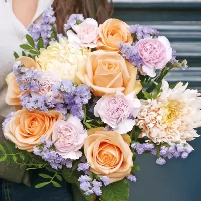 Bouquet de flores com rosas pêssego, cravos rosa e amarelos, flores lilases e folhas verdes.