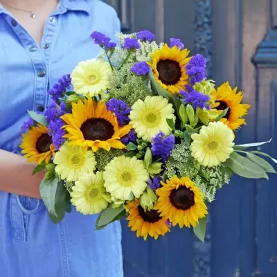 Bouquet de flores amarelas e roxas segurado por pessoa com vestido azul claro, fundo porta azul