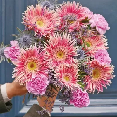 Bouquet de flores cor-de-rosa e azul-acinzentado segurado à frente de uma porta azul.