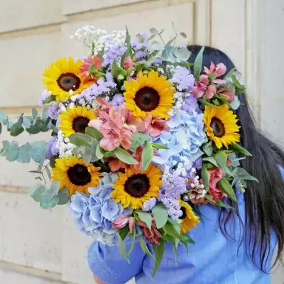 Arranjo colorido de flores com girassóis sendo segurado por pessoa de camisa azul