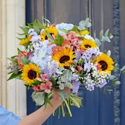 Bouquet colorido de flores variadas com girassóis e verdes segurado por mão com manga azul