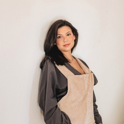 Mulher com macacão bege e camisa cinzenta encostada numa parede branca