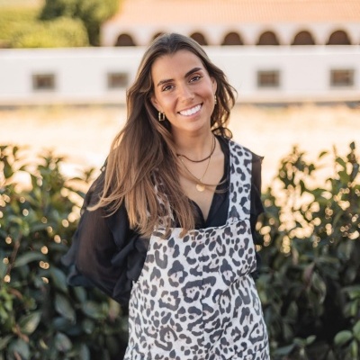 Mulher sorridente a usar macacão animal print preto e branco com blusa preta, em ambiente exterior com plantas e edifício ao fundo.