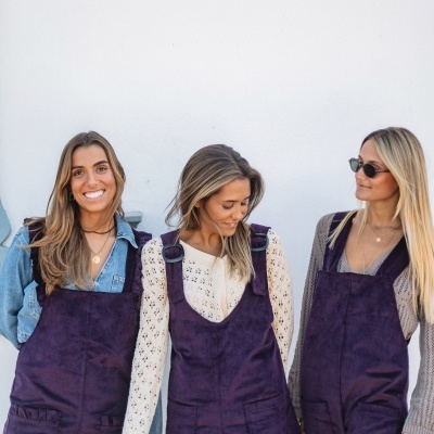 Três mulheres com jardineiras roxas de veludo e blusas diferentes em frente a uma parede branca