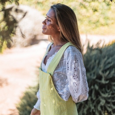 Mulher de lado com macacão amarelo claro e blusa branca bordada ao ar livre