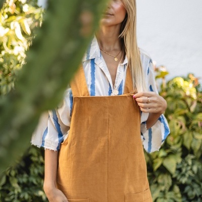 Mulher com macacão castanho e camisa riscada azul e branco junto a plantas e cacto