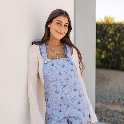 Mulher com macacão azul claro floral e camisola branca encostada numa parede exterior branca