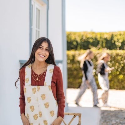 Mulher jovem com macacão floral e blusa vermelha junto a parede branca e cadeira de bambu