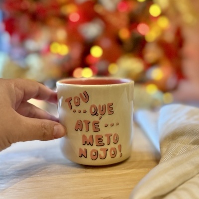 Caneca branca com interior vermelho e texto em rosa e preto segurada por uma mão em base de madeira