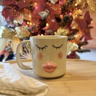 Caneca branca com rosto desenhado, sobre tábua de madeira e árvore de Natal ao fundo