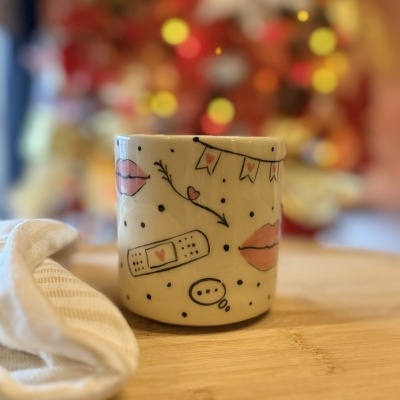 Caneca branca de cerâmica com desenhos de lábios, penso rápido com coração e bandeirinhas cor de rosa, sobre superfície de madeira clara