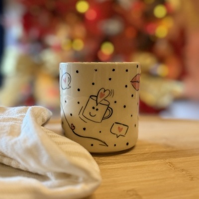 Caneca branca com desenhos de corações e pontos pretos sobre mesa de madeira com pano branco ao lado