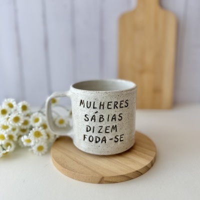 Caneca branca com texto preto sobre base de madeira e flores ao fundo