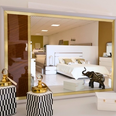 Espelho com moldura branca, detalhe castanho e dourado refletindo quarto moderno branco e bege