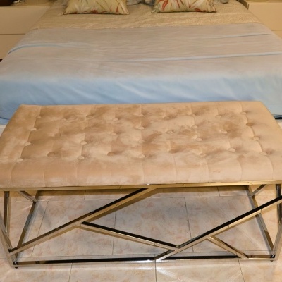 Banco retangular estofado bege com estrutura metálica prateada junto a cama com roupa azul clara