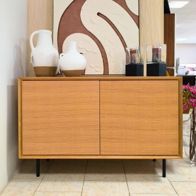 Aparador de madeira clara com duas portas em ambiente interior decorado com jarras cerâmicas e vaso de flores.