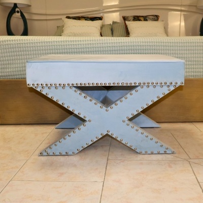 Banco azul claro estofado com tachas metálicas em base tipo X em chão de azulejos claro junto a cama.