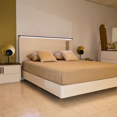 Cama branca com cabeceira alta e iluminação LED, roupa de cama bege com padrão xadrez e mesa de cabeceira com abajur circular dourado.