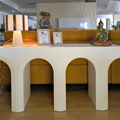 Mesa de consola branca com arcos, lâmpada com cúpula às riscas, molduras de fotos e estátua Buda, sofá amarelo ao fundo.
