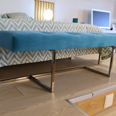 Banco azul de veludo com pernas metálicas cromadas junto a cama com capa de edredão geométrica