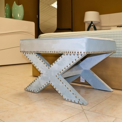 Banco azul claro em veludo com tachas douradas em forma de cruz, na sala com cama e cómoda branca.